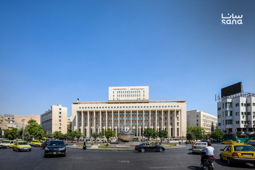 A central Damascus roundabout lined with taxis, framed by a colonnaded government building and a globe monument