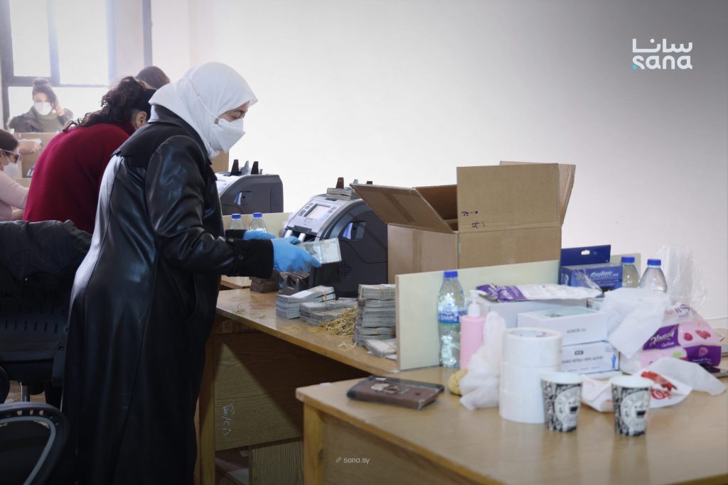 فنية تلبس قفازين وكمامة تعدّ أرزمة من الأوراق النقدية السورية القديمة بجانب آلتي عد نقود أثناء سحب الأوراق القديمة وإتلافها